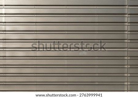 This image features a horizontal high-angle shot of a beige perforated metal surface with parallel ridges and visible mounting bolts.
The photograph captures a sophisticated industrial texture, showcasing a series of corrugated metal panels arranged in a seamless horizontal pattern. Each section of the material is meticulously detailed with a fine, repetitive micro-perforation that creates a subtle play of light and shadow across the surface. The neutral beige or champagne-toned finish lends a modern, architectural aesthetic to the composition, making it ideal for backgrounds related to engineering, urban design, or contemporary construction. Precisely spaced metallic bolts are visible along the recessed grooves, emphasizing the structural integrity and heavy-duty nature of the installation. The rhythmic repetition of the deep ridges provides a strong sense of linear perspective and geometric order. Soft, ambient lighting highlights the metallic sheen, revealing a slight iridescent quality in certain sections while maintaining a professional, clean look. This high-resolution texture exemplifies industrial minimalism, offering a versatile visual element for digital art, 3D mapping, or architectural visualization projects. The contrast between the dark shadows within the perforations and the reflective ridges adds depth and a tactile feel to the overall image. It serves as a perfect representation of durable building cladding or acoustic paneling used in modern commercial environments.
