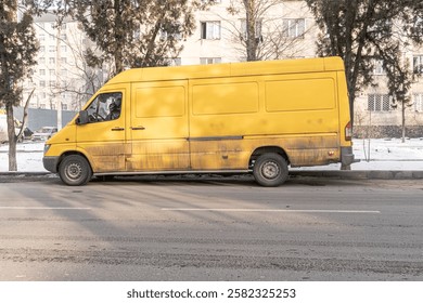 This image features a large yellow cargo van parked on the side of a city street during winter. The van appears worn and slightly dirty, with rust and grime along the lower panels. 