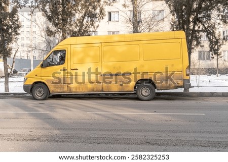 This image features a large yellow cargo van parked on the side of a city street during winter. The van appears worn and slightly dirty, with rust and grime along the lower panels. 