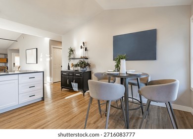 This image features a modern, open-concept kitchen and dining area, showcasing contemporary interior design trends such as warm wood flooring, neutral walls, and functional, stylish furniture. The dining set consists of a counter-height round table with a dark, likely metal or dark wood, base and top, paired with several grey velvet-upholstered counter-height bar stools. The use of upholstered seating and velvet fabric adds texture and an inviting warmth to the space The kitchen features sleek, white lower cabinets with dark countertops and simple black or dark pulls, contributing to the clean lines of the modern style. A prominent black sideboard with gold-tone handles sits against the wall, offering additional storage and a sophisticated design accent. This piece ties in with the growing trend of incorporating black accents and mixed metal finishes in modern kitchens. 