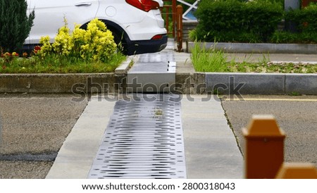 This image features a modern perforated metal drainage grate, central to urban water management and civil engineering design, wet from recent activity in a landscaped parking area. It highlights functional infrastructure detail, emphasizing efficient storm drain systems within a clean, contemporary urban environment. Ideal for illustrating sustainable city planning, public works projects, and environmental technology solutions in automotive or real estate development