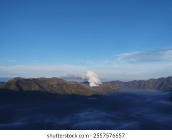 This image features a mountain with smoke rising from its peak. The base of the mountain is surrounded by clouds, while the sky above is clear and blue, creating a dramatic and natural atmosphere