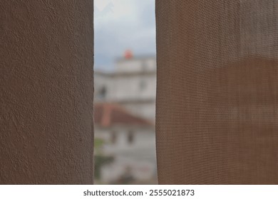 This image features a partial view of a curtain and a wall, creating a soft, textured foreground. The background is out of focus, showing a building with a red rooftop and a slightly cloudy sky