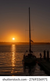 This image features a peaceful sunset over San Diego Bay, with a single-masted sailboat docked at a pier in the foreground. The warm, golden light of the setting sun creates a shimmering path across the water, silhouetting the boat's rigging and a lone figure on the dock against the vibrant sky.
