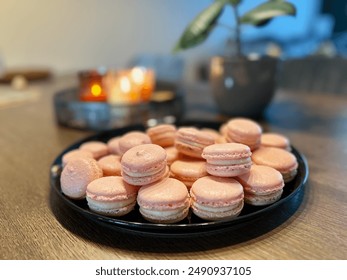 This image features a plate of pink home-made macarons on a wooden table, with a softly lit, cozy background that includes candles and a potted plant. - Powered by Shutterstock