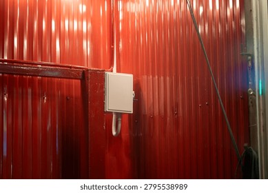 This image features a red corrugated metal wall indicating an industrial or construction setting, with an electrical box mounted and a hose extending along the wall.