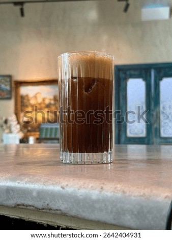 This image features a refreshing beverage, likely a cold coffee or a dark soda, served in a clear, ribbed glass with a substantial amount of foam on top.