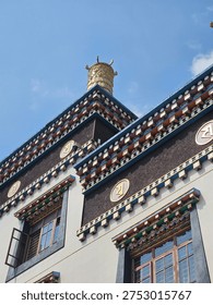 This image features the striking upper facade of a building designed in a traditional Tibetan Buddhist architectural style.

The structure is characterized by its clean white walls contrasted with dark, textured rectangular panels. These panels are decorated with large, golden circular medallions embossed with sacred scripts. Ornate, multi-layered eaves with colorful geometric patterns run horizontally across the building, leading up to a prominent, gold-plated victory banner (dhvaja) finial on the roof. The low-angle perspective highlights the intricate details against a clear,

vibrant blue sky.