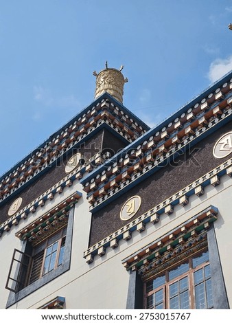 This image features the striking upper facade of a building designed in a traditional Tibetan Buddhist architectural style.

The structure is characterized by its clean white walls contrasted with dark, textured rectangular panels. These panels are decorated with large, golden circular medallions embossed with sacred scripts. Ornate, multi-layered eaves with colorful geometric patterns run horizontally across the building, leading up to a prominent, gold-plated victory banner (dhvaja) finial on the roof. The low-angle perspective highlights the intricate details against a clear,

vibrant blue sky.