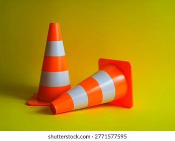 This image features two orange traffic cones, one standing upright and the other lying down, set against a bright yellow background. The cones have reflective white stripes and are commonly used for roadwork, construction, and other safety applications to divert traffic or indicate hazards.