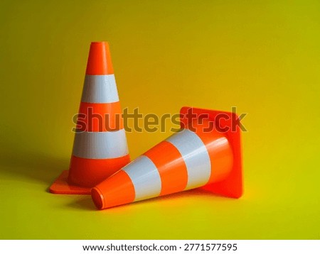 This image features two orange traffic cones, one standing upright and the other lying down, set against a bright yellow background. The cones have reflective white stripes and are commonly used for roadwork, construction, and other safety applications to divert traffic or indicate hazards.