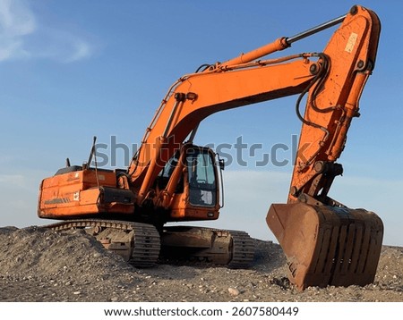 This image features a vibrant orange excavator perched atop a mound of dirt at an active construction site. The heavy-duty machine boasts a robust bucket and hydraulic arm