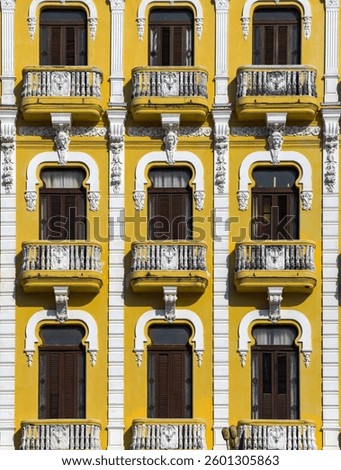 This image features a vibrant yellow building facade adorned with ornate white architectural details, including decorative balconies, intricate carvings, and symmetrically framed windows.cuba .havana