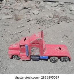 This image features a worn, red plastic toy semi-truck sitting outdoors on dusty, dry ground.