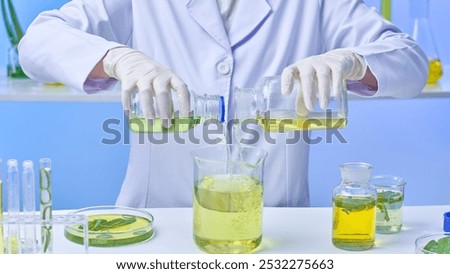 In this image of a modern laboratory, samples of aloe vera and other flasks are arranged around a scientist who is pouring a pale yellow solution from two tiny flasks into a huge pale yellow beaker.