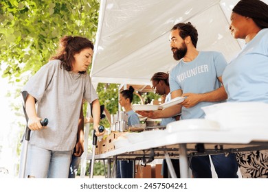 In this image, multiracial volunteers providing hunger relief support to caucasian woman on crutches. Voluntary individuals distribute free food, offer shelter, and assistance to poor and disabled.