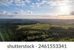 This image offers a breathtaking aerial perspective of the Hautes Fagnes region, capturing the vast expanse of rolling hills and dense forests under a sunlit sky. The late afternoon sun casts dramatic