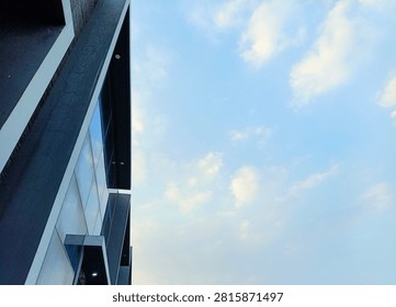 This image presents a compelling composition that juxtaposes modern architecture with an expansive sky. On the left side, a segment of a contemporary building facade is prominently featured. The structure boasts a strong geometric design, primarily in dark gray or black, accented by sharp white or light gray lines and panels that create a striking contrast. Several glass panels reflect the soft blue and white of the sky, adding a dynamic element and depth to the building's surface. Subtle cracks are visible on some of the darker sections of the wall, hinting at texture and age despite the modern aesthetic. The perspective is an upward, low-angle shot, emphasizing the building's height and the interplay of its vertical and diagonal lines. Recessed lights are also visible on the underside of some overhangs.

The majority of the right side of the image is dominated by a bright, clear blue sky dotted with a few wispy white clouds. This vast expanse of sky provides a serene negative space that beautifully contrasts with the rigid, angular lines of the man-made structure. The lighting indicates a bright daytime setting, casting an even glow over the scene. The overall impression is one of minimalist and contemporary aesthetics, with a strong focus on form, lines, and the interaction between urban design and natural elements.