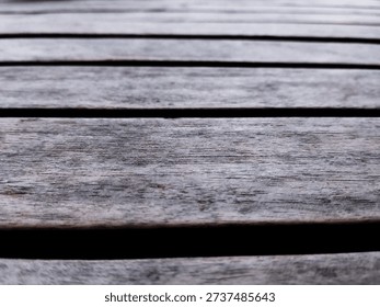 This image presents a compelling study of texture, perspective, and the natural passage of time, focusing on a weathered wooden surface. The composition is dominated by horizontal planks of aged timber, arranged in a parallel fashion that creates a rhythmic, linear pattern. The camera angle is low and angled, utilizing a shallow depth of field that draws the viewer’s eye from the sharp, tactile details of the foreground into a soft, hazy blur toward the top of the frame. This bokeh effect lends a sense of infinite scale to the surface, transforming a simple wooden deck or table into a vast, rugged landscape.
​The wood itself tells a story of exposure to the elements. Its color palette is a sophisticated mix of muted greys, earthy browns, and subtle charcoal tones, characteristic of timber that has been bleached by the sun and worn by rain. Every plank is etched with a complex web of fine lines, grooves, and grain patterns, showcasing the organic complexity of the material. The dark, narrow gaps between the slats provide a sharp tonal contrast, emphasizing the structural repetition of the surface. There is an inherent 