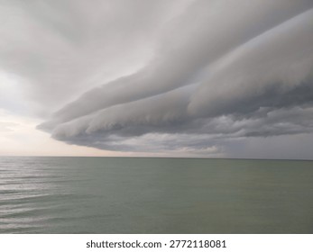 This image presents a dramatic seascape under an overcast sky. A vast expanse of calm, pale-green ocean stretches toward the horizon, where it meets a heavy formation of layered storm clouds. The clouds appear thick and sweeping, creating a striking, almost wave-like shape in the sky that suggests an approaching storm. Subtle tones of gray, blue, and faint pink near the horizon add depth and contrast to the scene. In the distance, a small boat is visible as a dark silhouette floating quietly on the water, emphasizing the scale and emptiness of the surrounding environment. The overall mood of the image is tranquil yet slightly tense, capturing the quiet stillness before a possible storm.