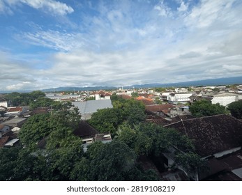 This image presents a panoramic view of a town surrounded by lush greenery under a vast, dramatic sky. The foreground is filled with dense trees and houses with red-tiled roofs, creating a peaceful residential landscape. In the distance, more buildings stretch toward a range of mountains that forms a beautiful natural backdrop. Above, the sky is covered with a mixture of white and gray clouds, allowing patches of blue to shine through and adding depth to the scenery. The combination of urban development, abundant vegetation, and the scenic mountain view creates a calm and refreshing atmosphere, showcasing the beauty of the landscape.