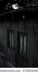 This image presents a somber, high-angle view of a weathered wooden building, likely a shed or small rural house.

Structure and Materials
Wooden Siding: The walls are constructed from vertical wooden planks that are aged and darkened, showing significant weathering and a rough, splintered texture.

Green-Trimmed Windows: Two small, side-by-side windows are set into the wall. They feature worn green frames and white or light-colored panes that appear dirty and aged.

Clay Tile Roof: The top of the image shows a portion of a traditional clay tile roof. The tiles are covered in patches of vibrant green moss, suggesting a damp environment and a long period of exposure to the elements.

Lighting and Atmosphere
Low Light: The image is quite dark, with heavy shadows obscuring many details of the wooden grain and the area beneath the eaves.

Mood: The combination of the dark lighting, weathered wood, and overgrown moss creates a quiet, melancholic, or perhaps abandoned atmosphere.

Background: In the very top of the frame, dark silhouettes of tree leaves are visible against a pale, overcast sky.