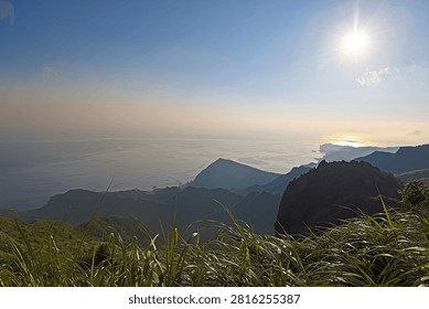 This image presents a stunning panoramic view from Teapot Mountain in Taiwan, looking toward the Pacific Ocean. The high-angle perspective captures layered mountain ridges sloping down to the coastline, where the sea meets a hazy sky. Bright sunlight reflects beautifully off the water, highlighting the majestic landscape.