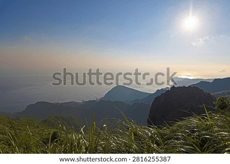 This image presents a stunning panoramic view from Teapot Mountain in Taiwan, looking toward the Pacific Ocean. The high-angle perspective captures layered mountain ridges sloping down to the coastline, where the sea meets a hazy sky. Bright sunlight reflects beautifully off the water, highlighting the majestic landscape.