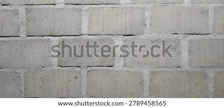 This image presents a close‑up view of a sturdy wall constructed from uniformly grey concrete blocks or cinder blocks. The rectangular blocks are laid neatly in a running‑bond pattern, bonded together by lighter grey mortar joints that create clear, straight lines across the surface. Each block has a coarse, porous, matte texture typical of raw concrete—simple, solid, and unadorned. The overall appearance is clean, symmetrical, durable, and minimalist, highlighting the raw, utilitarian strength and uniform structure of masonry work.