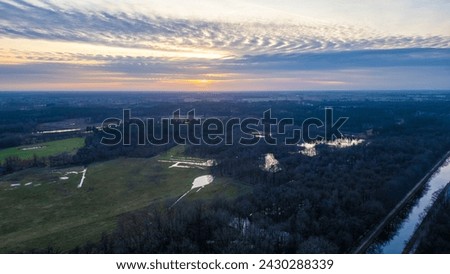 This image provides a breathtaking aerial view of a tranquil landscape at sunset. The sun dips towards the horizon, its rays filtering through the clouds, casting a warm glow over the land. Water