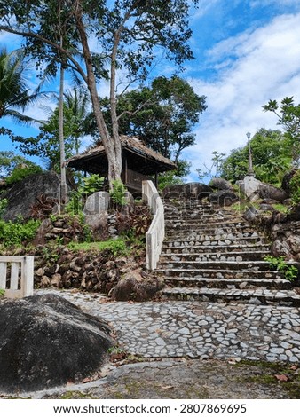 This image showcases a beautiful, rustic tropical garden path leading up a hillside. A wide flight of stone steps, built with embedded rocks and flanked by a white concrete handrail, curves upwards toward a traditional wooden gazebo featuring a thatched roof. The surrounding landscape is lush and natural, dotted with large, smooth granite boulders, tropical trees (including coconut palms), and leafy green plants. Above the garden, a bright blue sky filled with wispy white clouds adds a serene and refreshing atmosphere to the entire scene.