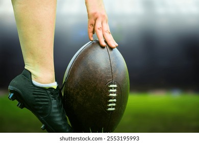 This image showcases a close-up of a rugby or American football player's hand placing a ball on the ground, with a cleated foot supporting it. The setting suggests a game or practice environment. - Powered by Shutterstock