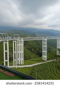 This image showcases a scenic elevated walkway extending over a lush green landscape,The structure is supported by tall white pillars with geometric patterns, giving it a modern and artistic look. 