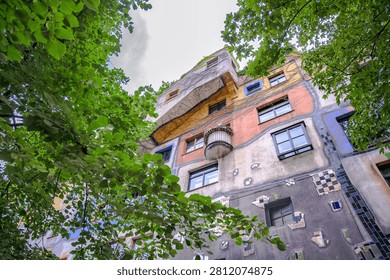 This image showcases the vibrant facade of the Hundertwasserhaus building in Austria standing tall amidst lush green trees and a cloudy sky with colorful patches of br