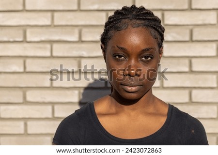This image showcases a young African woman standing confidently in front of a white brick wall. The subject is centered in the frame, with her hair neatly styled in cornrows that add a textural