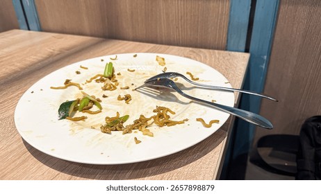 This image shows an almost empty white plate with leftover fried noodles, scattered crumbs, and a fork and spoon, indicating a recently finished meal.