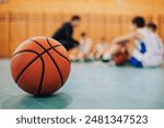 This image shows a close-up of a basketball on a gym floor, with blurred kids training in the background, highlighting the focus on the ball and the ongoing sport activity