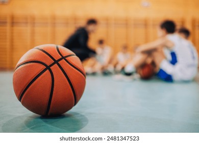 This image shows a close-up of a basketball on a gym floor, with blurred kids training in the background, highlighting the focus on the ball and the ongoing sport activity - Powered by Shutterstock