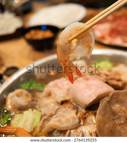 This image shows a close-up of a cooked shrimp held by chopsticks over a steaming Thai BBQ grill (Mu Kratha), surrounded by sliced pork and vegetables.