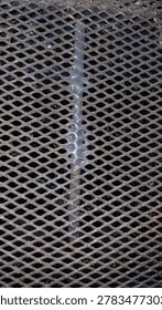 This image shows a close-up of an expanded metal mesh surface with a rugged industrial appearance. The metal sheet features a repeating diamond-shaped pattern created through the expansion process, giving it a strong yet lightweight structure. A visible welded seam runs vertically through the center, while the slightly rusted and weathered texture adds a raw, metallic character. The dark background behind the mesh enhances the depth and geometric design, making it suitable for construction, fencing, ventilation panels, or industrial décor applications.