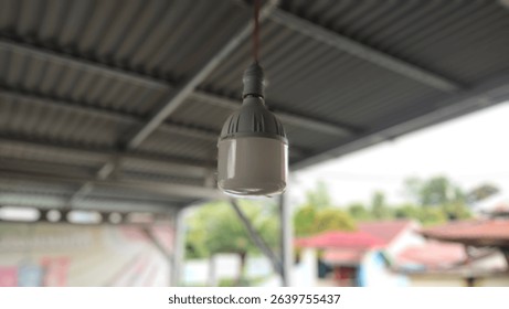This image shows a close-up of an LED light bulb hanging from the ceiling of a covered outdoor area. The bulb is attached to a red electrical wire and has a white and gray plastic casing.