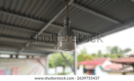 This image shows a close-up of an LED light bulb hanging from the ceiling of a covered outdoor area. The bulb is attached to a red electrical wire and has a white and gray plastic casing.
