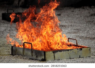 This image shows a controlled fire burning inside a rectangular metal tray placed outdoors on a gravel or gravel like ground surface.