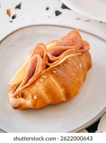 На этом изображении показан вкусный сэндвич с круассаном из ветчины и сыра. Он украшен красиво золотистым, шелушащимся круассаном, увенчанным морскими соляными хлопьями и наполненным тонко нарезанной ветчиной и сыром, скорее всего 