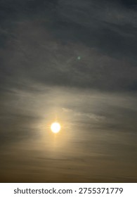 This image shows a dramatic sky with a bright celestial body, likely the sun or moon, piercing through dense, moody clouds above a dark silhouette of a town skyline.
The scene depicts a 