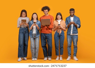 This image shows a group of five young adults standing in a row against a yellow background. They are all looking down at their devices, which include a tablet, a smartphone, and a laptop - Powered by Shutterstock