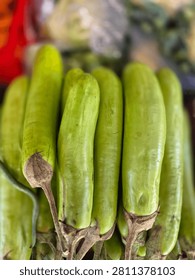 This image shows a group of fresh green eggplants displayed at a market. The eggplants are long and smooth, with shiny light-green skin and brown stems. They are arranged closely together, creating an attractive and natural appearance. The blurred background highlights the vegetables and gives the photo a vibrant and fresh atmosphere.