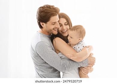 This image shows a happy family of three, a man, a woman, and their baby, standing close together in a white interior setting. The man and woman are smiling at the baby - Powered by Shutterstock