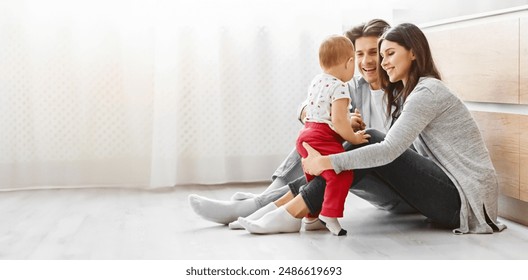 This image shows a happy family, a mother, father, and baby, playing together on a light wood floor in their living room. The parents are seated on the floor and the baby is seated in the mothers lap