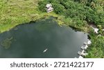 This image shows a high-angle drone view of a calm, dark lake surrounded by lush green tropical vegetation. A small boat glides across the water, with white lakeside cabins and a hidden building visible